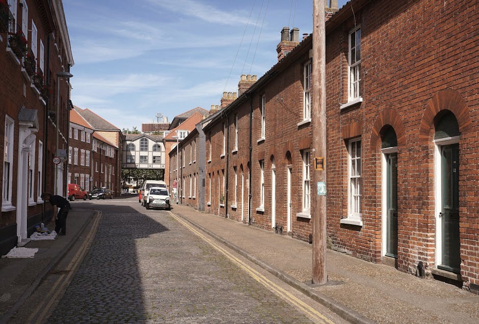 A narrow cobblestone street in Guildford with brick terraced houses on the right and parked cars further along. A person is seen on the left side wearing dark clothing, handling packing materials such